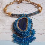 Pendentif Corsica avec un cabochon : une Agate Bleue brodée avec des perles nacrées Bleu Lapis en cristal à laquelle s’ajoute des rocailles saphir et travertin entrelacées pour former une dentelle